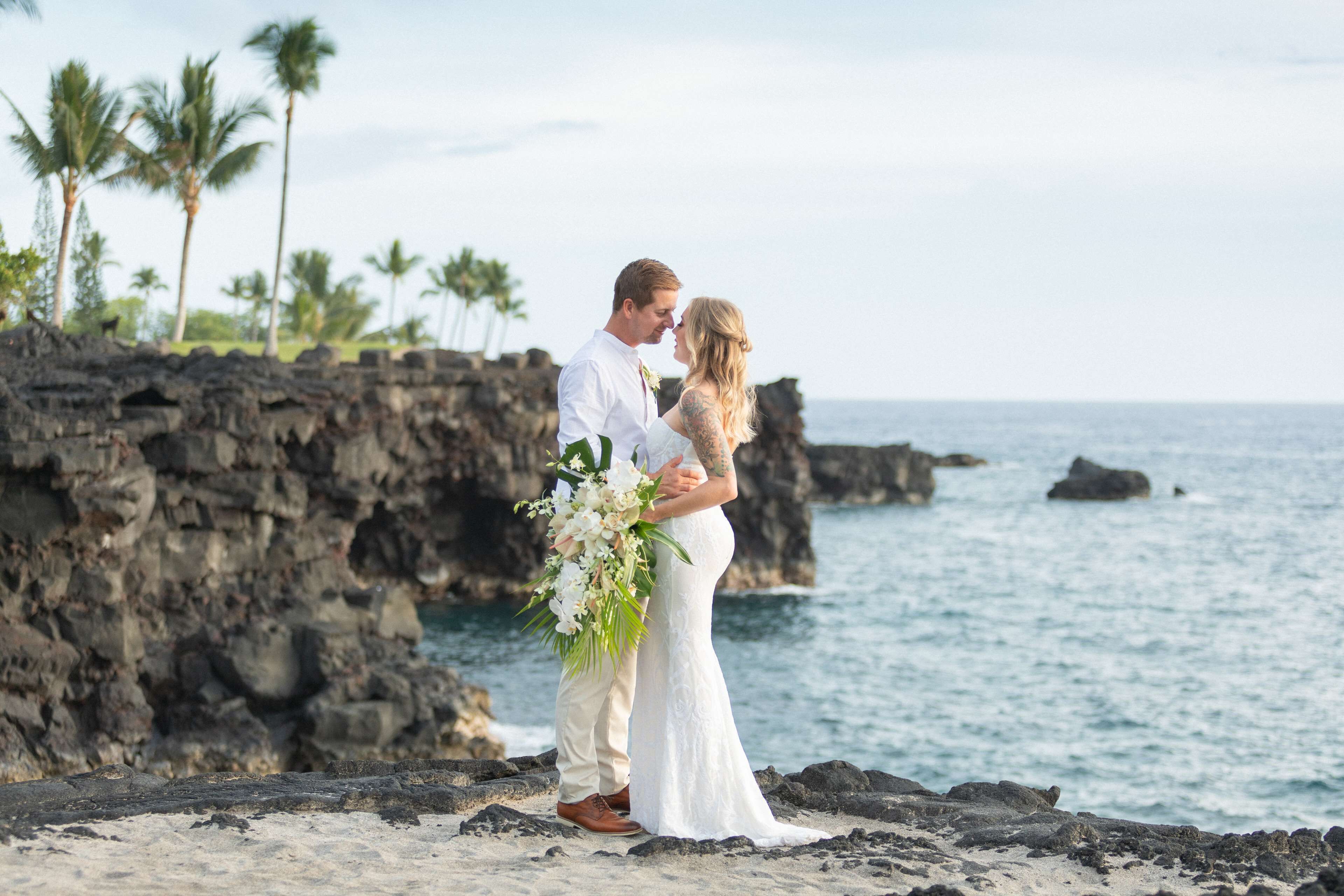 A couple embracing on a rocky beach with palm trees and ocean in the background.