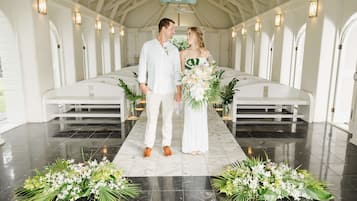 Ein Paar steht in einer Kapelle mit weißen Kirchenbänken und floralen Dekorationen auf dem Boden.