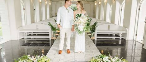 A couple standing in a chapel with white pews and floral arrangements on the floor.