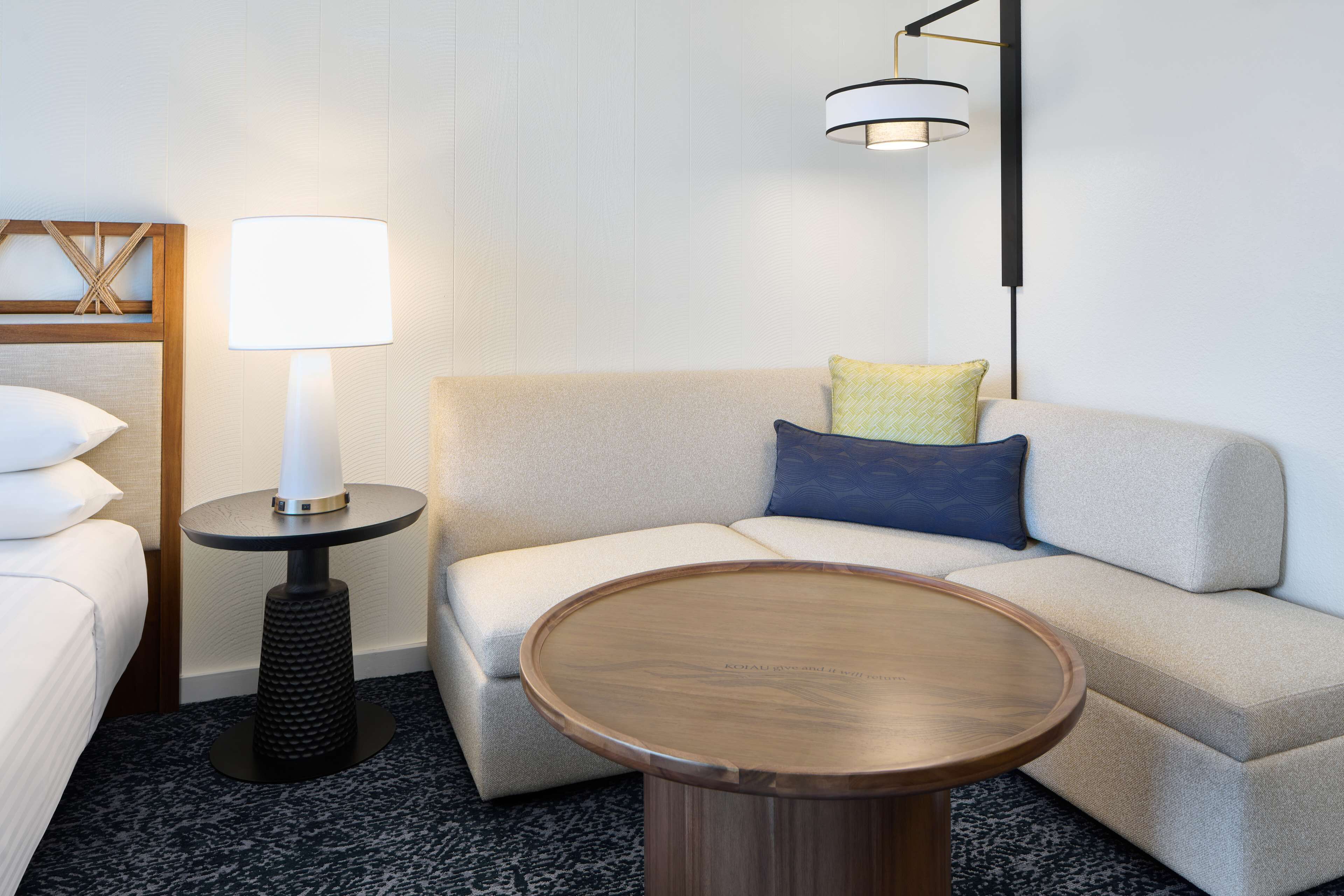 A modern hotel room with a sofa, a round wooden coffee table, a lamp, and a bed.