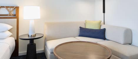 A modern hotel room with a sofa, a round wooden coffee table, a lamp, and a bed.