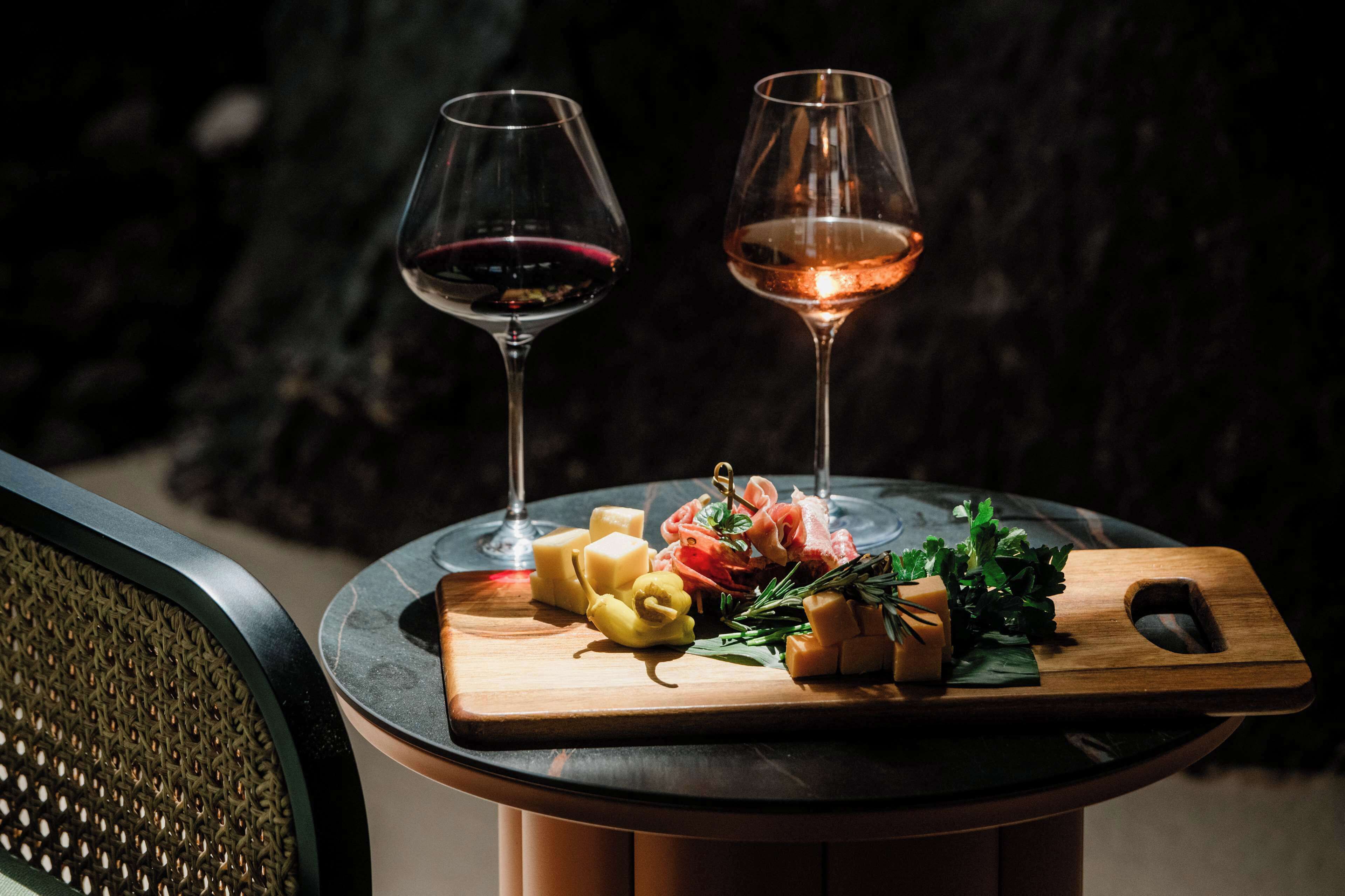 Una mesa con dos copas de vino, una tabla de madera con queso, verduras y hierbas.