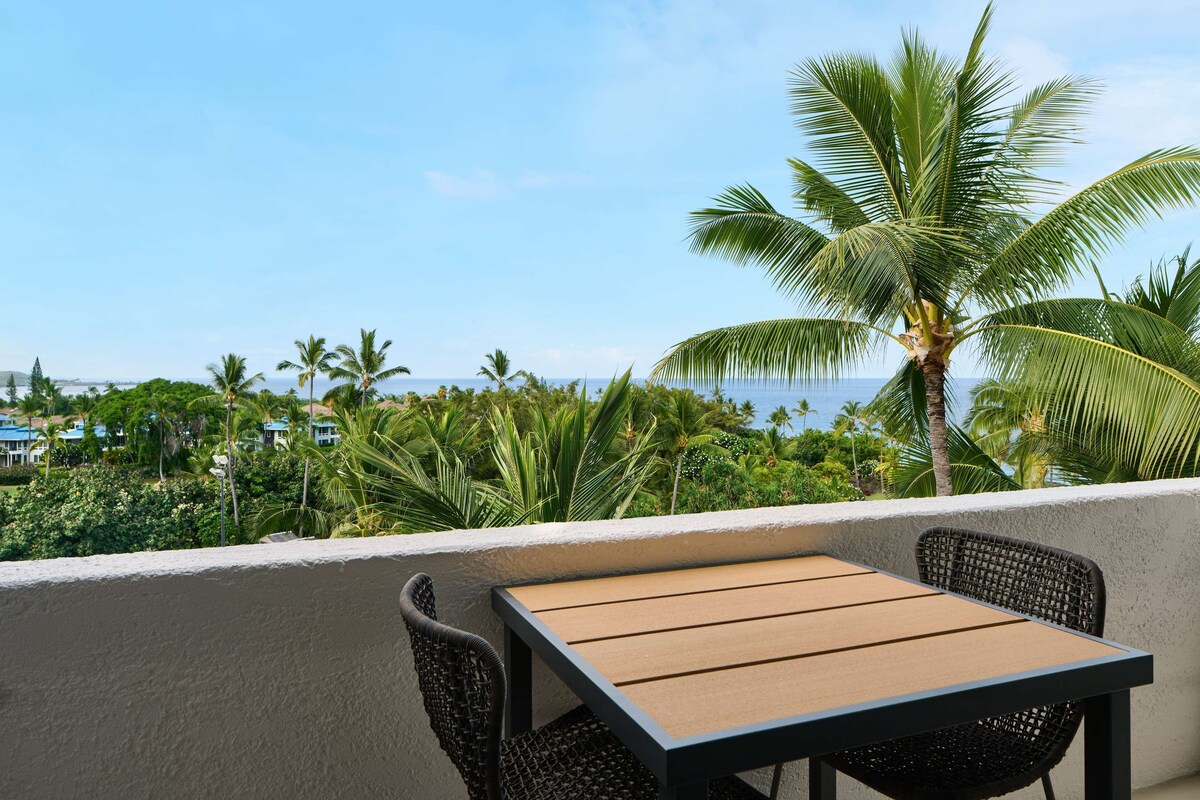 Un balcon avec une table et des chaises, donnant sur une zone côtière bordée de palmiers et de bâtiments.