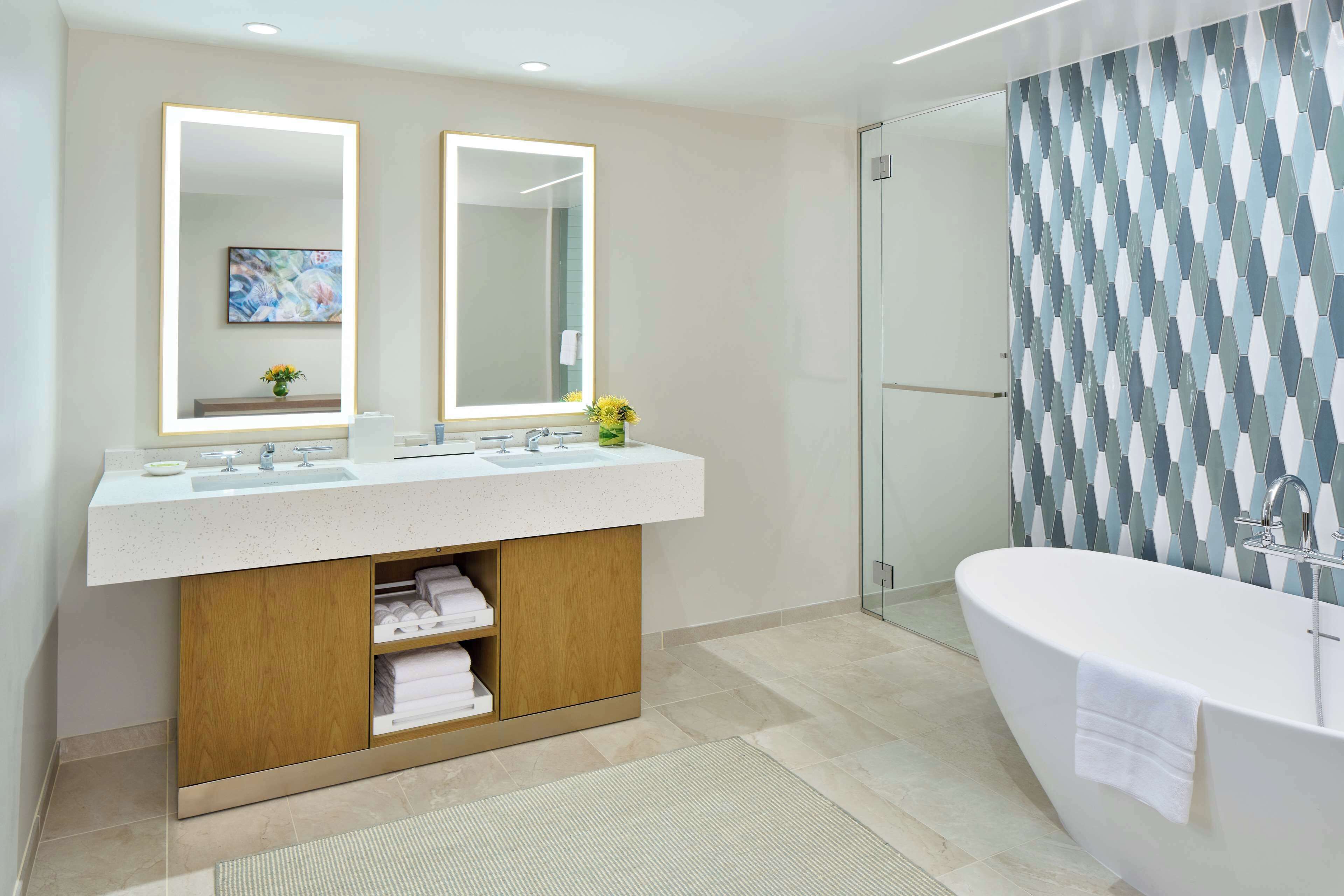 Une salle de bain moderne avec une baignoire sur pieds, un meuble-lavabo double doté de miroirs rétroéclairés et un mur de douche orné d’un motif géométrique.