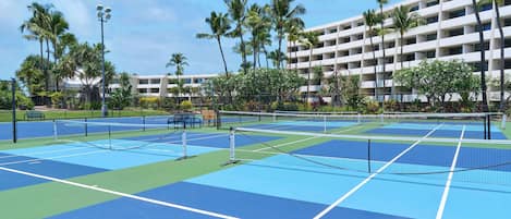 An outdoor tennis court with multiple courts, surrounded by palm trees and a multi-story building in the background.