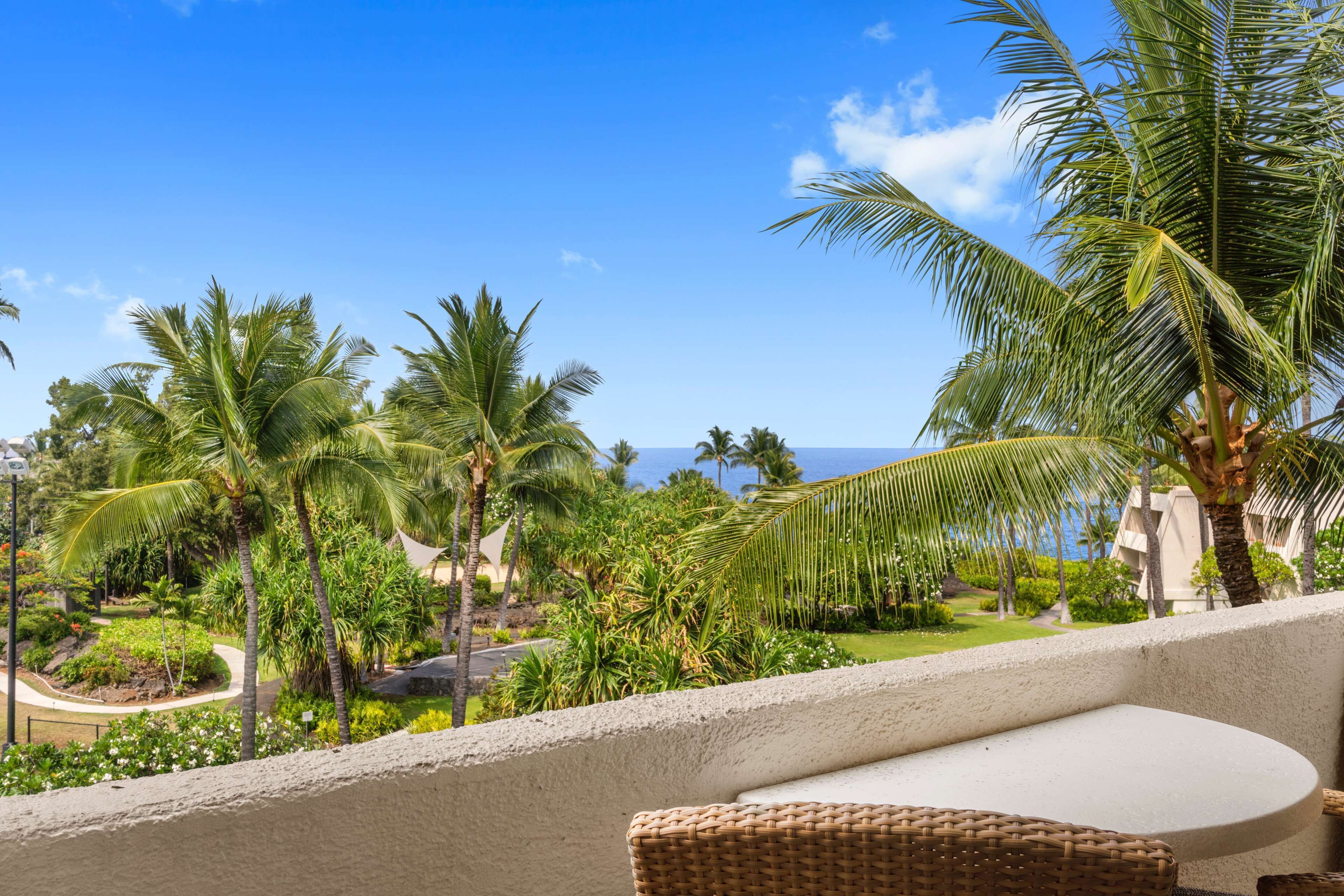 A balcony with wicker furniture, overlooking a tropical garden with palm trees and a clear blue sky.