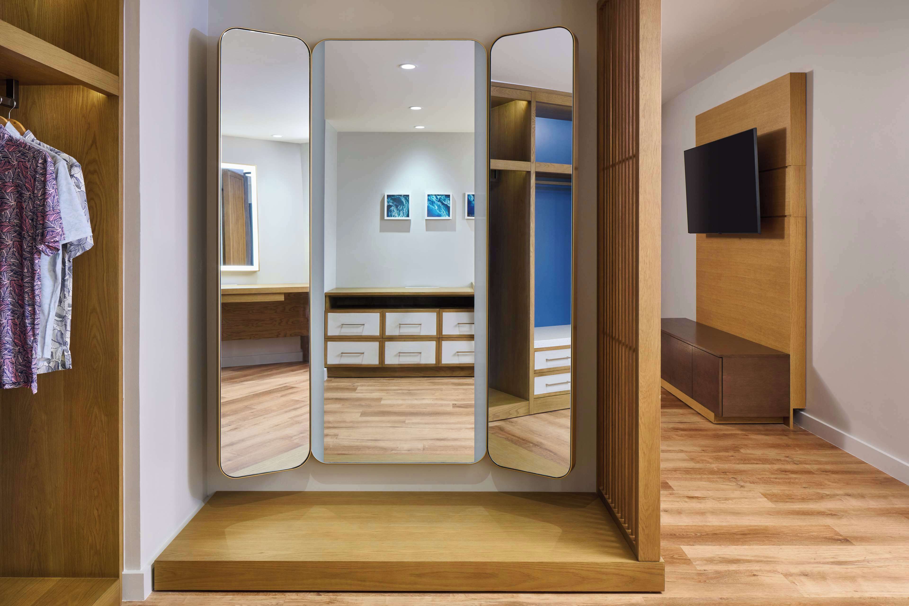 Una habitación con un armario de madera, un espejo, una cama con cabecero azul y un televisor fijado a la pared.