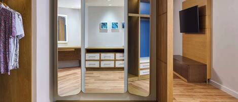 A room with a wooden wardrobe, a mirror, a bed with a blue headboard, and a TV mounted on the wall.