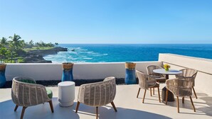 A terrace with wicker furniture and a round table, overlooking a coastal view with the ocean and palm trees.