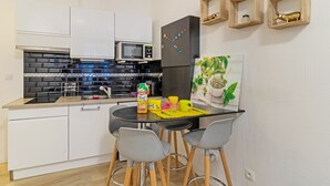 Una cocina compacta con armarios blancos, un salpicadero negro y un microondas. Hay un refrigerador negro con imanes de colores, una alfombrilla amarilla sobre la mesa y dos sillas grises.
