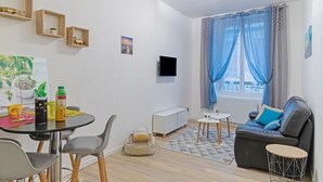 Una sala de estar moderna con un sofá, una mesa de comedor con sillas, un pequeño armario blanco, un televisor montado en la pared y una ventana con cortinas azules.