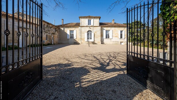 A courtyard with a building featuring a tiled roof and white shutters, surrounded by a gravel path and metal gates. - Domaine du chauvet (Ambares-et-Lagrave)