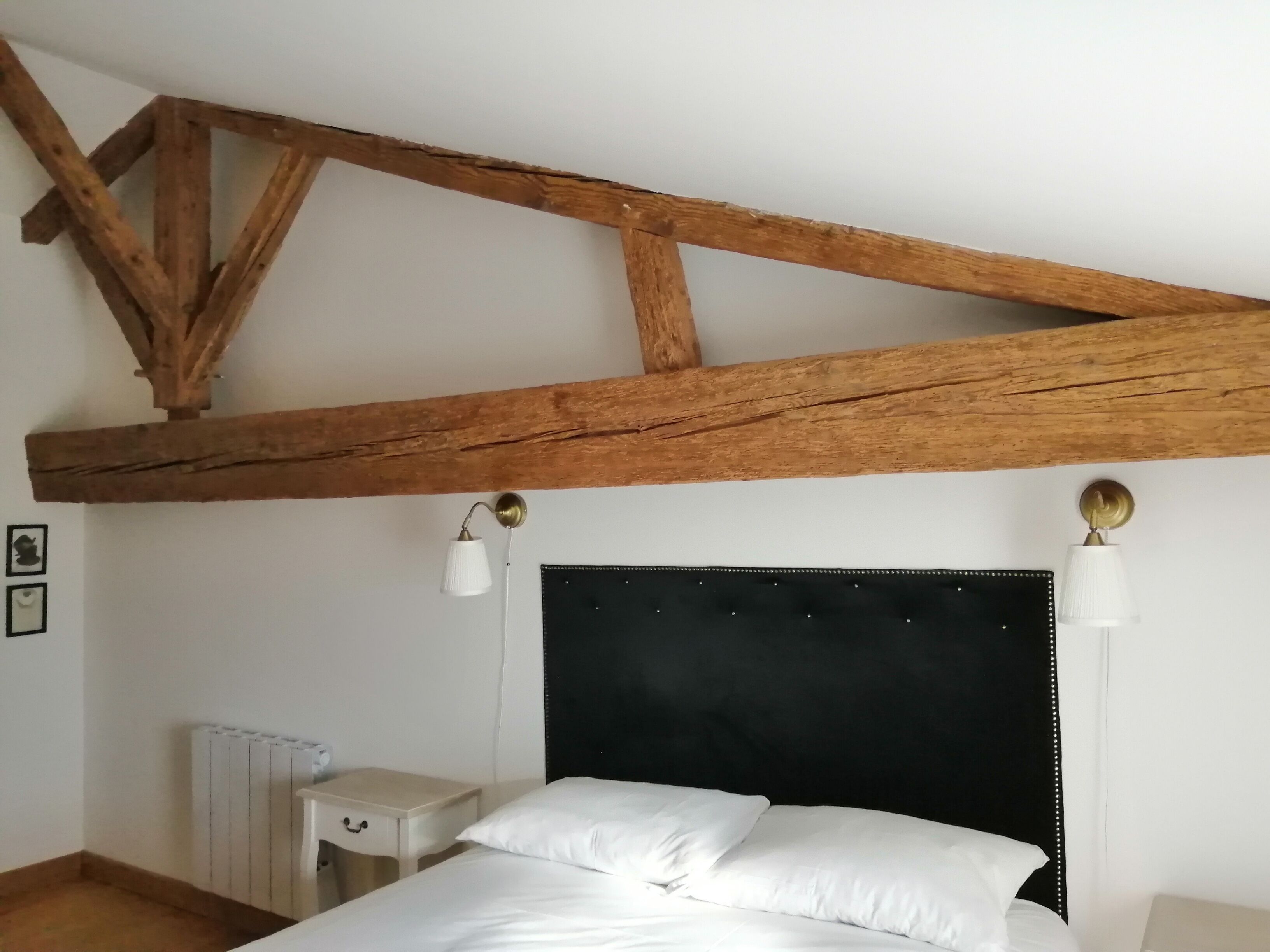 Un dormitorio con vigas de madera a la vista, cabecero negro, ropa de cama blanca, mesitas de noche y lámparas fijadas a la pared.