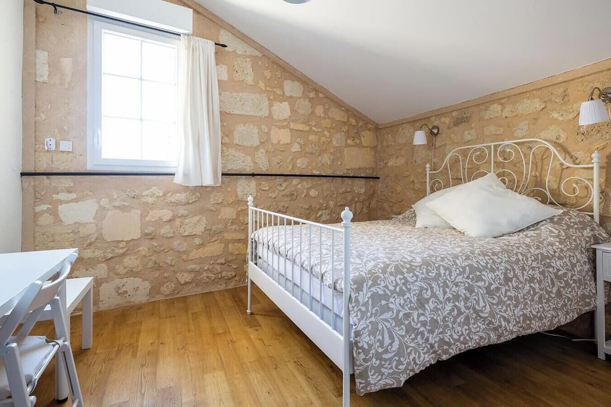 Un dormitorio con pared de piedra, cama con ropa de cama estampada, un escritorio blanco y una ventana con cortinas.
