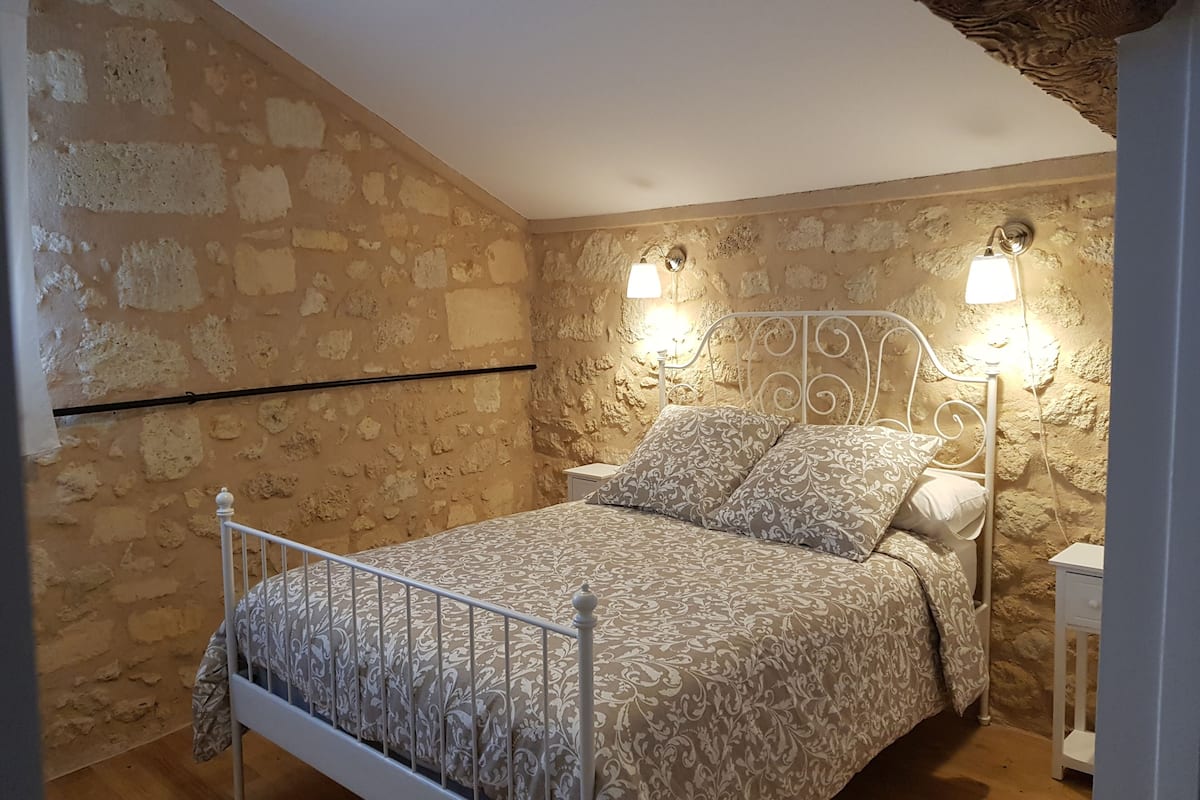 Un dormitorio con pared de piedra, estructura de cama metálica, ropa de cama estampada, dos lámparas de noche y suelo de madera.