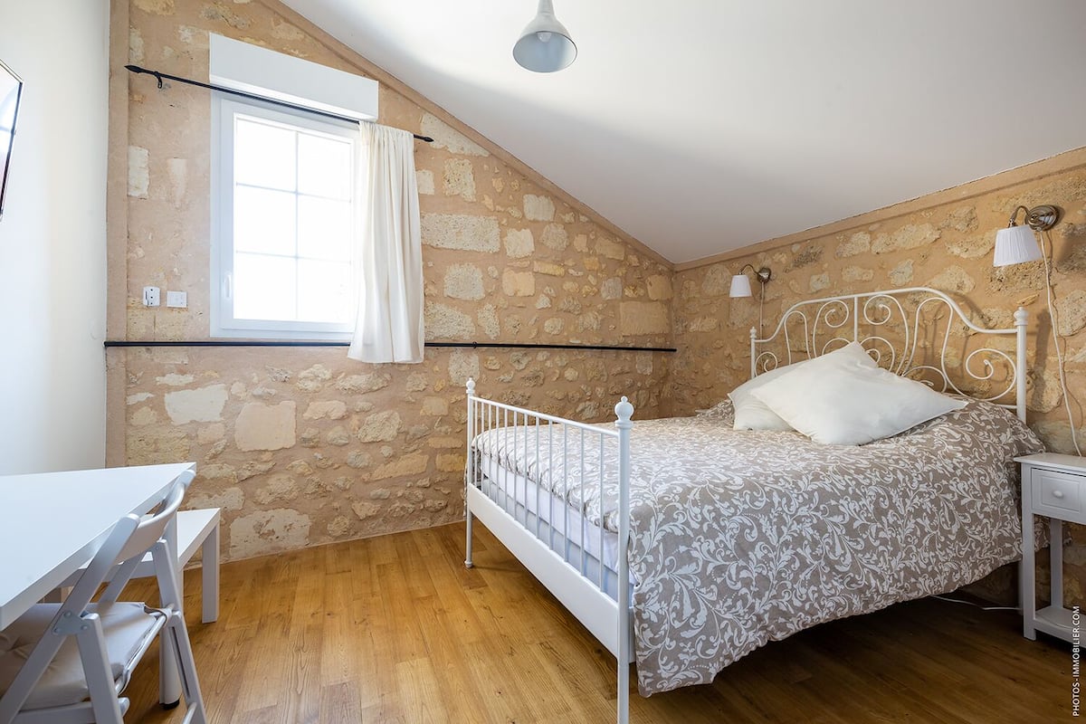 Un dormitorio con pared de piedra, una cama con cabecera blanca, una mesita de noche, un escritorio y una ventana con cortina.