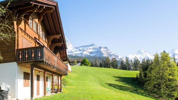 Un chalet traditionnel en bois, doté d’un balcon, qui se dresse sur un fond de montagnes enneigées et d’une colline verdoyante.
