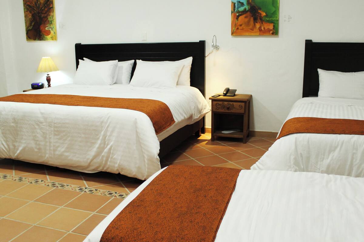 Ein Hotelzimmer mit zwei Betten, jeweils mit weißer Bettwäsche und braunen Kopfteilen. Zwischen den Betten befindet sich ein Nachttisch mit Lampe und Telefon. Der Raum hat Fliesenboden und Kunstwerke an den Wänden.