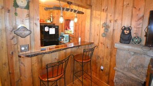 A rustic wooden interior with a kitchen area featuring a black refrigerator, a wooden bar counter, two metal bar stools, and a stone fireplace. - Little Mosser-Awesome Views! Very Quiet! (Estes Park)