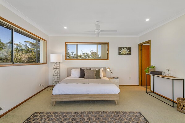 A bedroom with a large bed, a ceiling fan, a bedside table with a lamp, a window with a view of trees, and a small table with a plant.