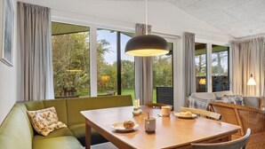 A dining area with a wooden table, green sofa, wicker chairs, and large windows offering a view of the outdoors. - Amazing home in Hjerm with house a panoramic view (Hjerm)