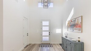 A hallway with a light wooden floor, a gray cabinet with a mirror and a plate, a door with frosted glass panels, and a painting on the wall. - Hideaway On The 2nd Green (St. George)