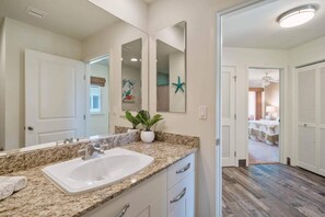 Une salle de bain avec un comptoir en granit, un lavabo blanc et un grand miroir. On trouve une plante en pot sur le comptoir et une décoration en forme d’étoile de mer sur le mur.