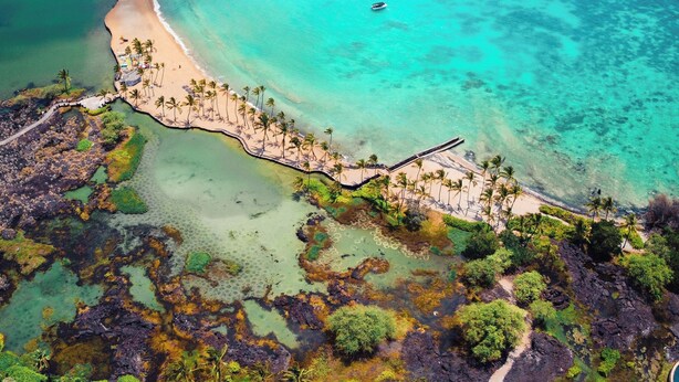 An aerial view of a coastal area with clear turquoise waters, a sandy beach, and a boat anchored near the shore.