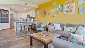 A modern living room with a gray sofa, a wooden coffee table, and a kitchen area with white cabinets and a bar stool.