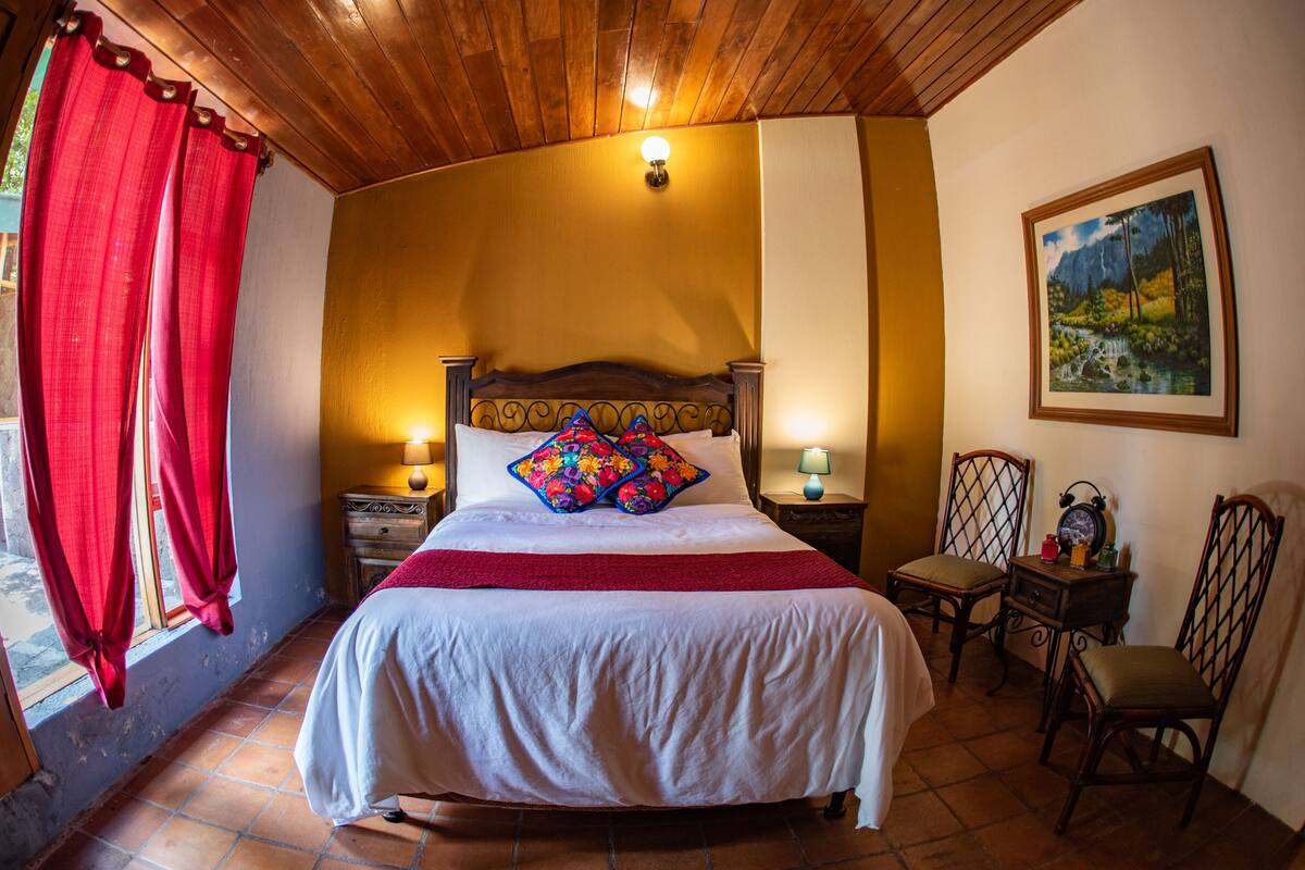 Een slaapkamer met een houten plafond, een bed met een rode deken, twee nachtlampjes, een schilderij aan de muur en een raam met rode gordijnen.