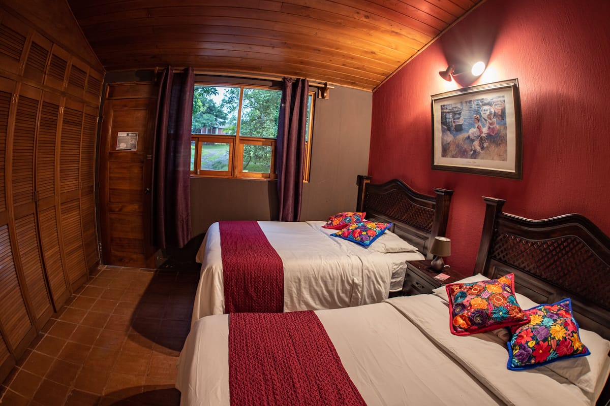 Een kamer met twee bedden, houten hoofdeinden, een raam met gordijnen, een ingelijst kunstwerk aan de muur en een houten deur.