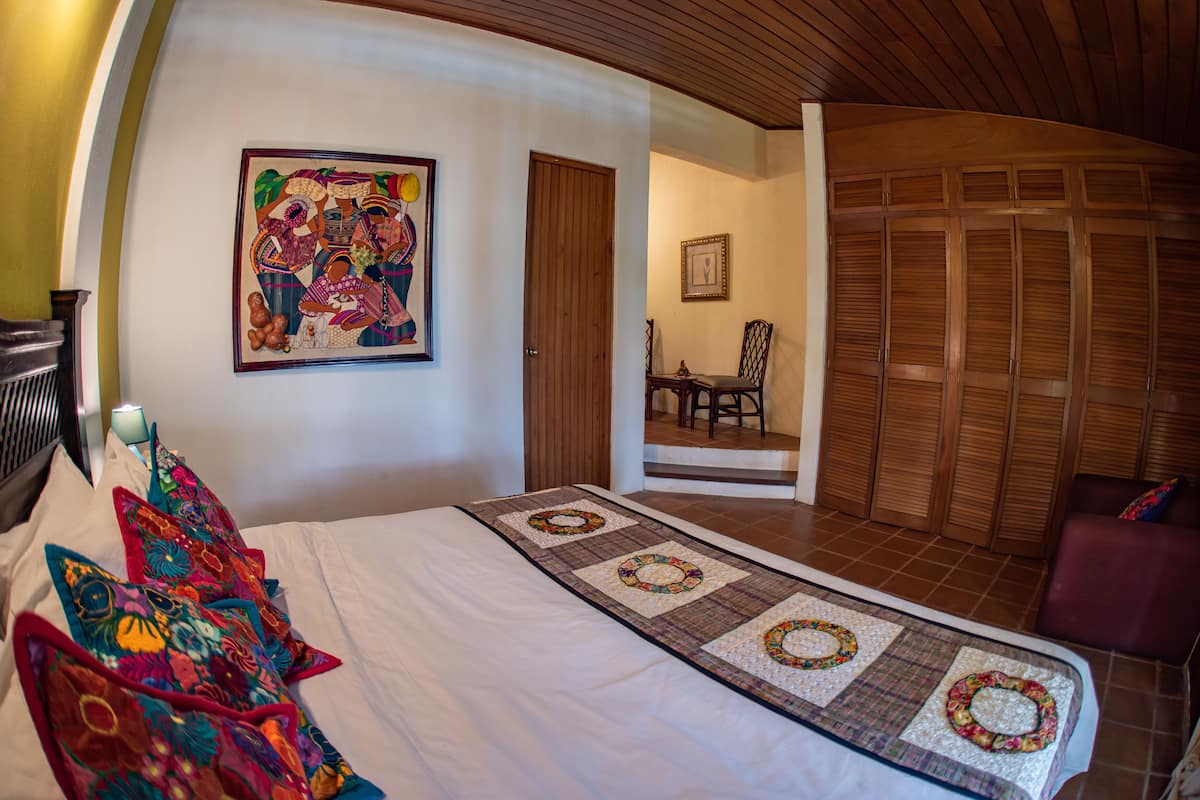 Een hotelkamer met een bed, een sprei, een kleurrijk schilderij, een houten deur en een stoel.
