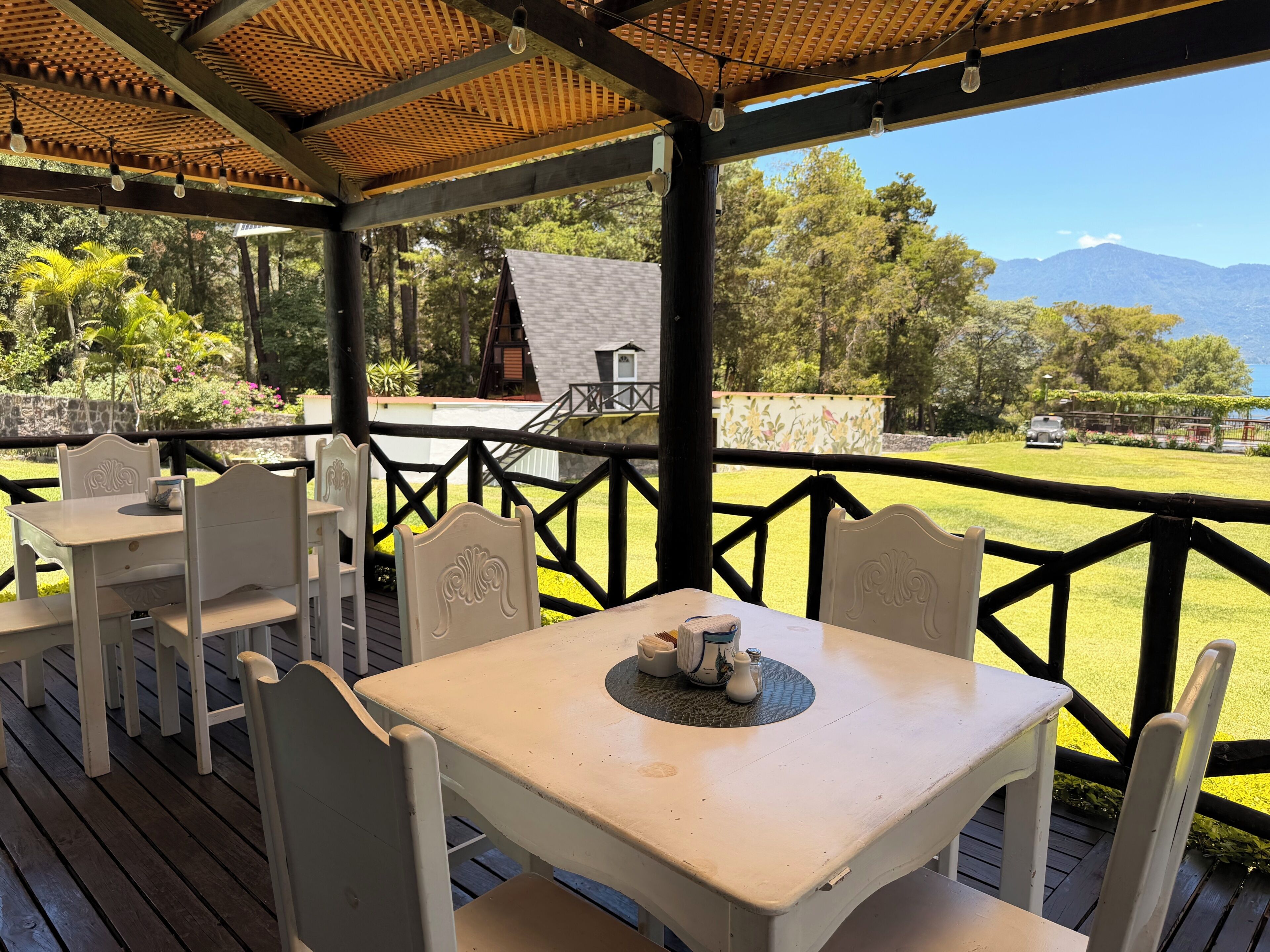 A covered outdoor seating area with tables and chairs, overlooking a grassy landscape and a mountain in the distance.