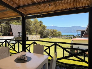 A covered outdoor seating area with a table and chairs, overlooking a grassy landscape and a body of water with mountains in the distance. - Hotel Los Olivos (Santiago Atitlán)