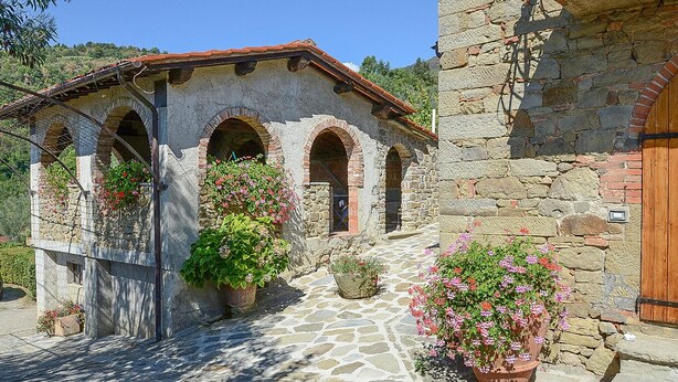 A stone building with arched windows and a wooden door. There are potted plants with flowers in front of the windows. - La Casella- La Terrazza (Castelfranco di Sopra)