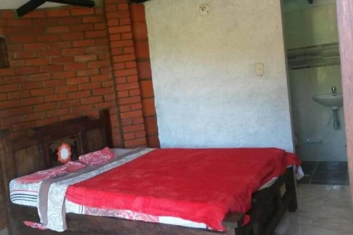 A room with a brick wall, a wooden bed with a red blanket, and a white wall with a sink in the background.