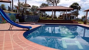 A swimming pool with a slide, surrounded by a tiled patio and a gazebo.