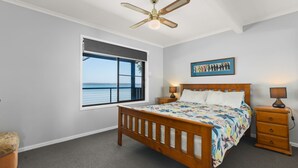 Ein Schlafzimmer mit großem Fenster, einem Bett mit farbenfroher Tagesdecke, zwei Nachttischen mit Lampen, einem Deckenventilator und einem gerahmten Kunstwerk an der Wand.