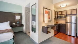 Habitación de hotel con cama, mesita de noche, lámpara, espejo y una kitchenette equipada con refrigerador y microondas.