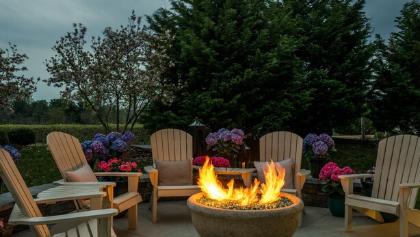 Un patio con sillas Adirondack, un hogar de piedra con llamas y flores en macetas.