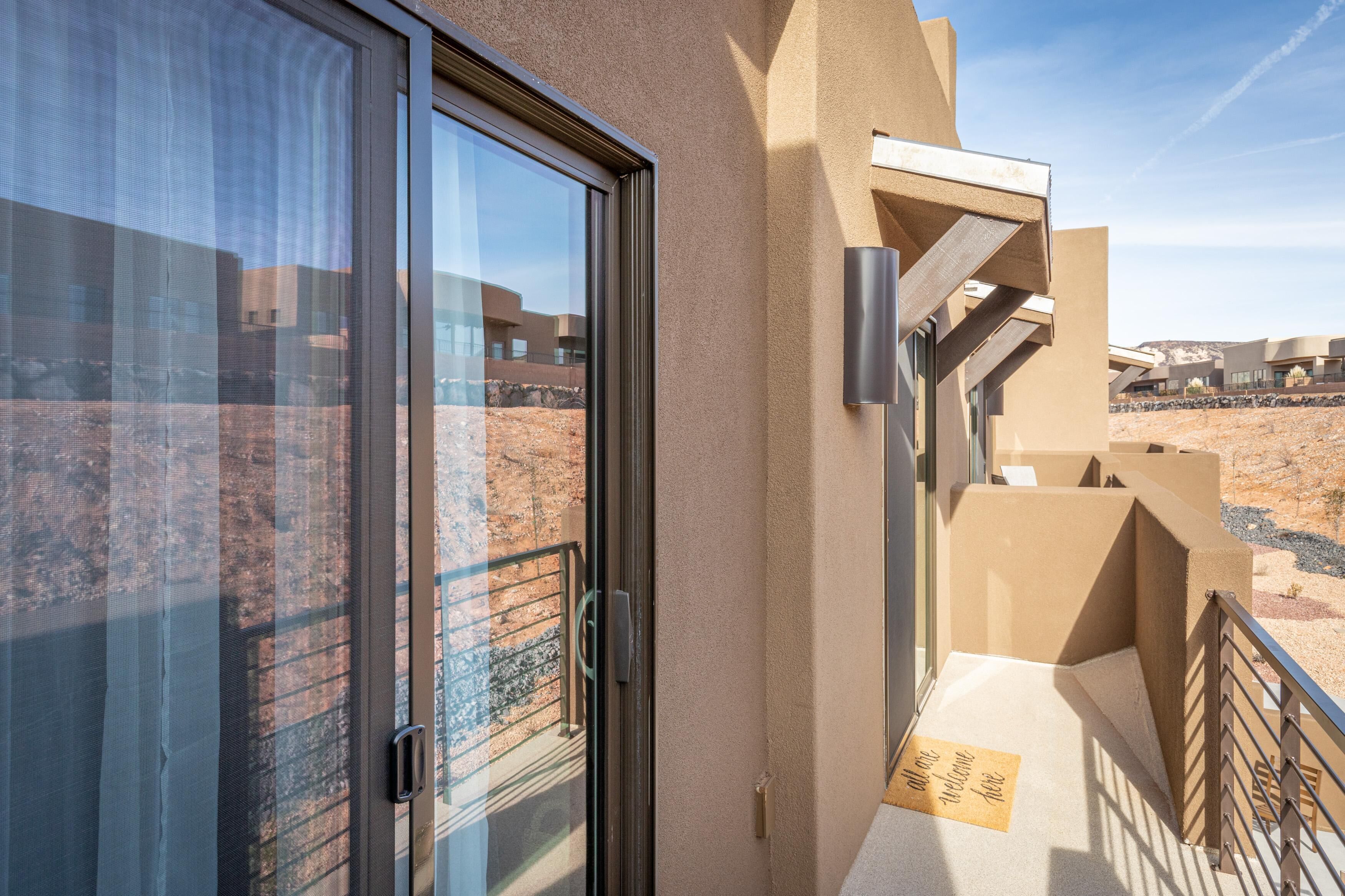 A modern hotel room with a large sliding glass door, a balcony with a metal railing, and a view of a desert landscape.