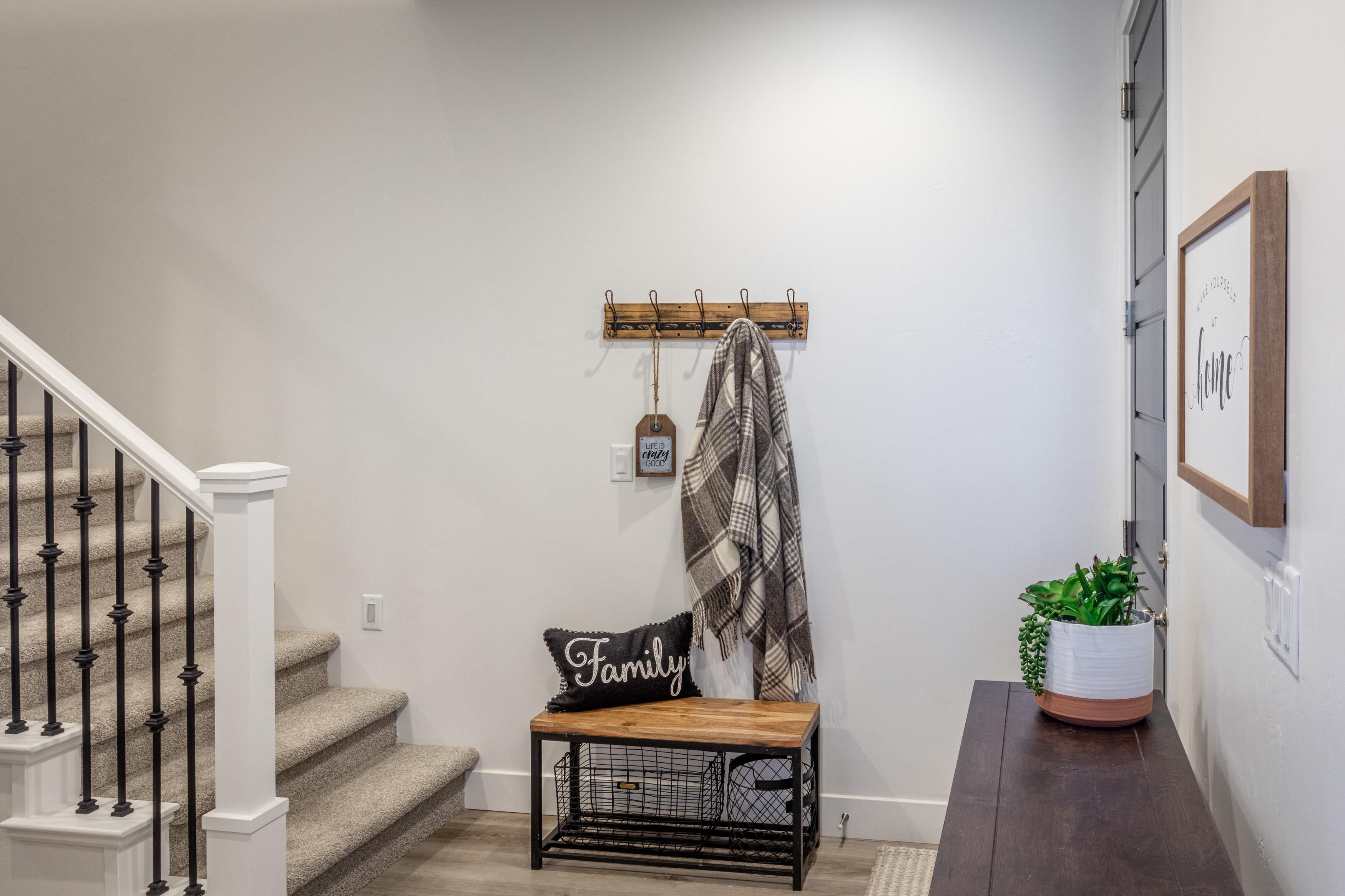 Un escalier avec une rampe blanche, un banc agrémenté d’un coussin orné du mot « Famille », un porte-manteau avec une écharpe à carreaux, et une plante en pot.