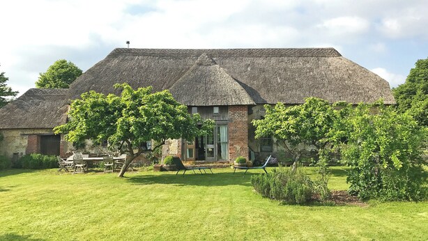 Une maison à toit de chaume, avec une entrée en briques, entourée d’une pelouse bien entretenue et d’arbres.