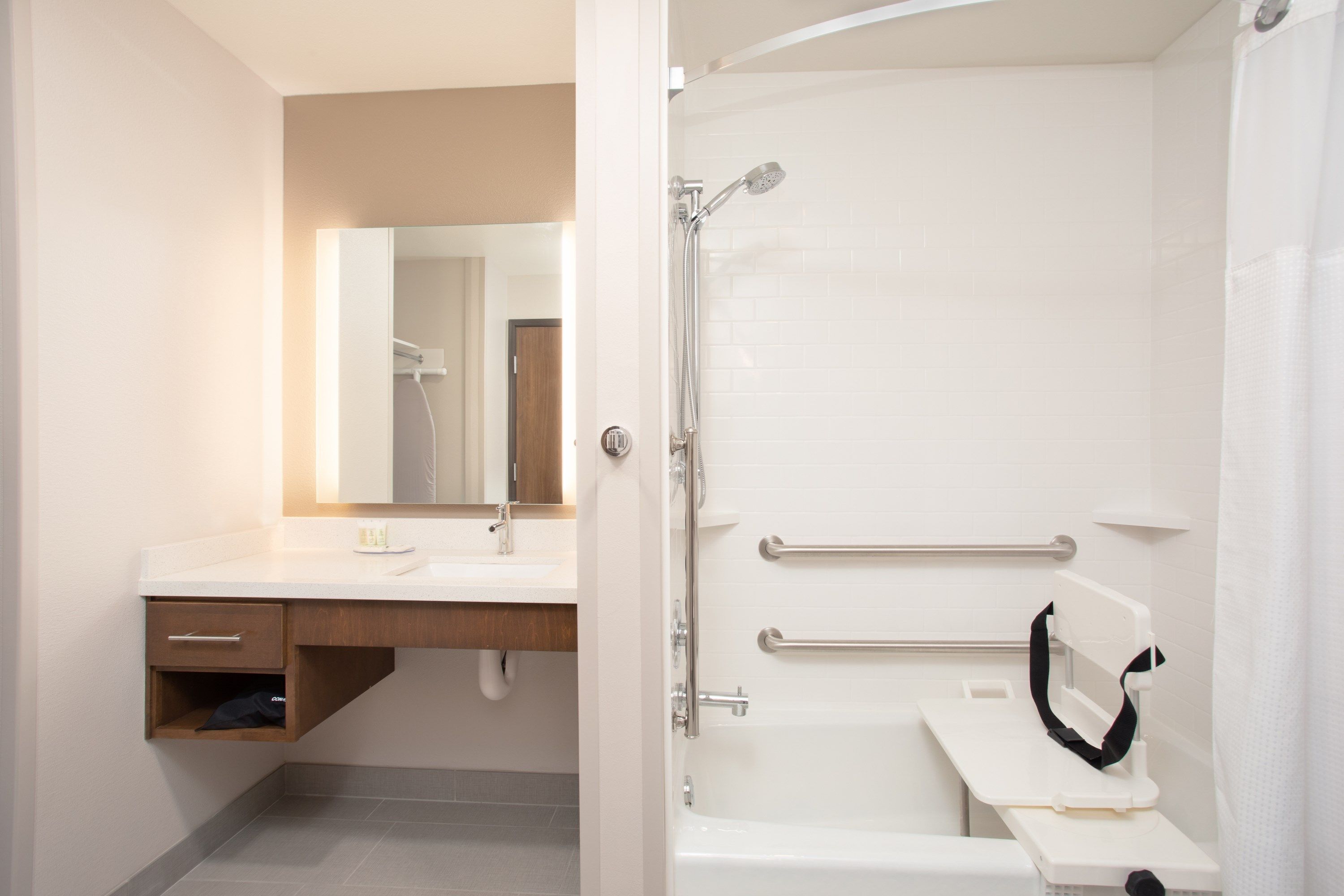 A modern bathroom with a white sink, mirror, and shower. It includes a grab bar for safety and a towel rack.