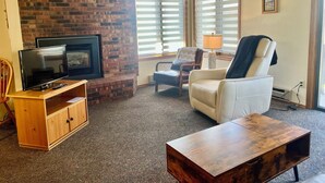 A living room with a brick fireplace, a television, a leather armchair, and a wooden coffee table.