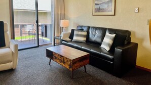 A living room with a black leather sofa, a wooden coffee table, a white armchair, and a large window with a view of a deck.