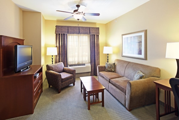Habitación de hotel con televisión de pantalla plana, ventilador de techo, sofá, una silla, mesa de centro y lámpara.