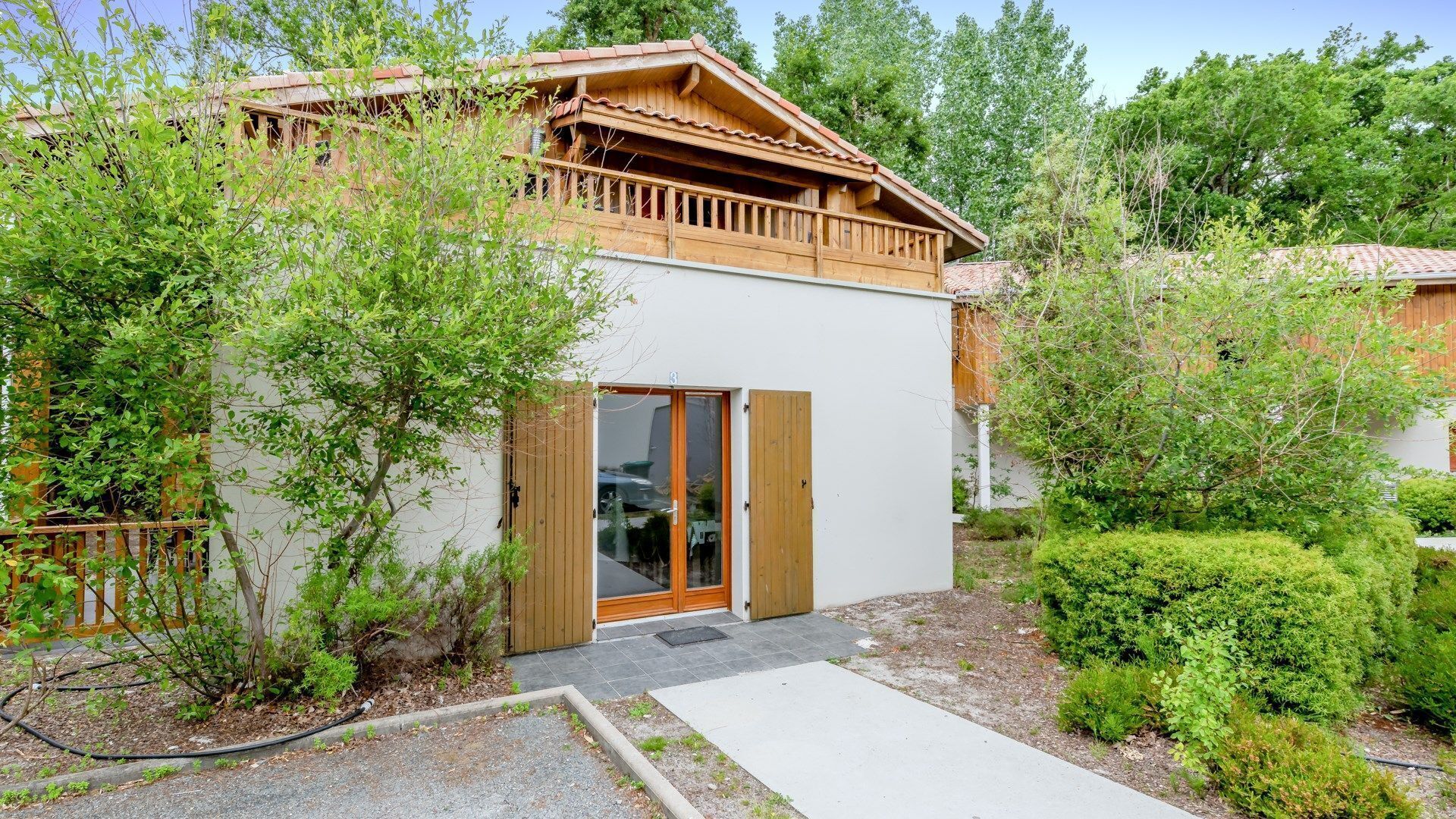 Une petite maison en bois d’un seul étage, dotée d’un balcon, entourée d’une végétation luxuriante et accessible par une allée en gravier.