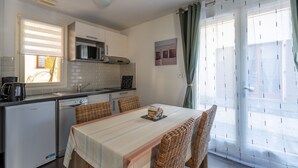 A compact kitchen with a microwave, sink, and a small refrigerator. There is a dining table with four wicker chairs, and a window with curtains. - Appart charmant + accès piscine (Lacanau)