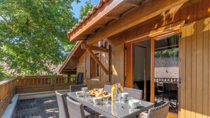 Une terrasse extérieure en bois, dotée d’une table à plateau de verre et de chaises en osier, entourée d’une clôture en bois et d’arbres.